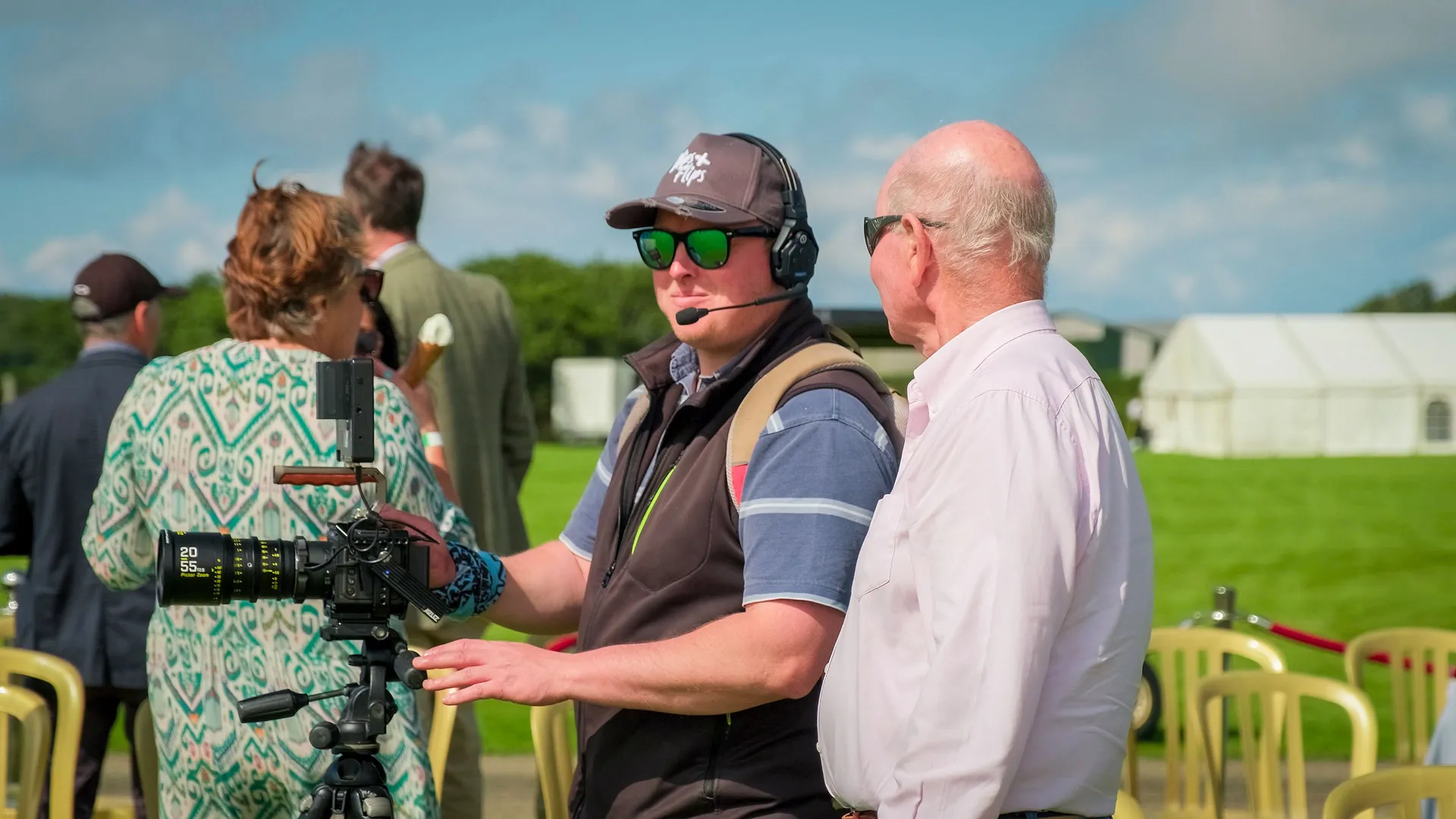 Two men stand outdoors; one operates a camera, wearing a headset and sunglasses. The other, in a white shirt, watches. Green field and tents in the background.